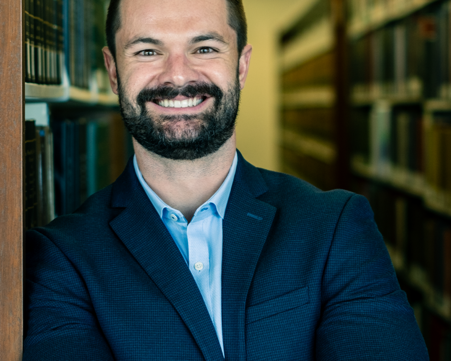 Copywriting Interview Profile picture: Photograph of a smiling man pictured in a library wearing a dark suit.