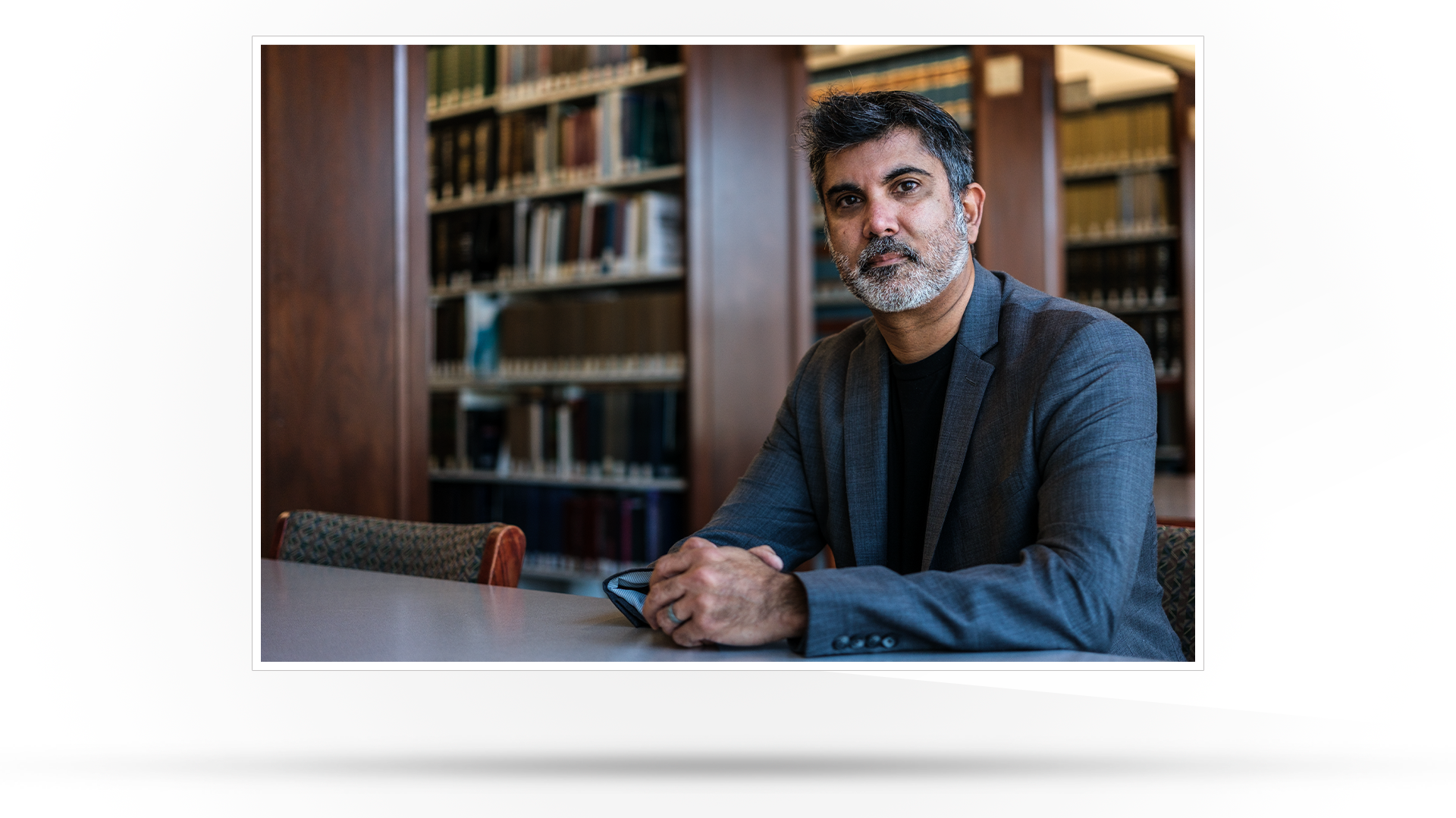 Copywriting Interview Profile picture: Photograph of a male member of faculty pictured in a library wearing a dark suit.